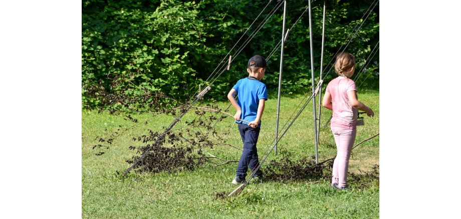 Zwei Kinder sind zu sehen. Diese stehen ind er grünen Natur und ziehen hinter sich einen großen Ast mit vertrockneten Blättern her. 