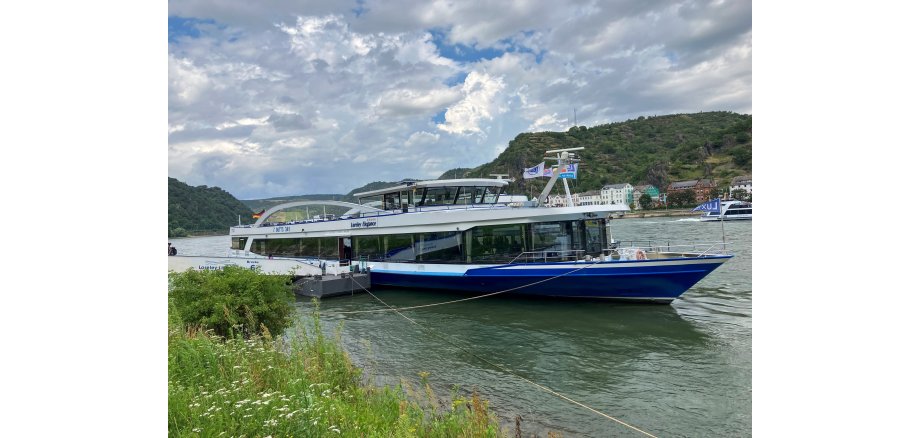 Auf dem Bild erkennt man ein Schiff. Dieses liegt an einem grünen Rhein-Ufer. Das Schiff ist blau und weiß. Am Himmel erkennt man weiße Wolken.