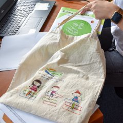 Man sieht, wie eine Frau in einen hellbraunen Stoffbeutel ein Schulbuch einpackt. Auf diesem Beutel ist das Logo der Verbandsgemeinde Nieder-Olm abgebildet